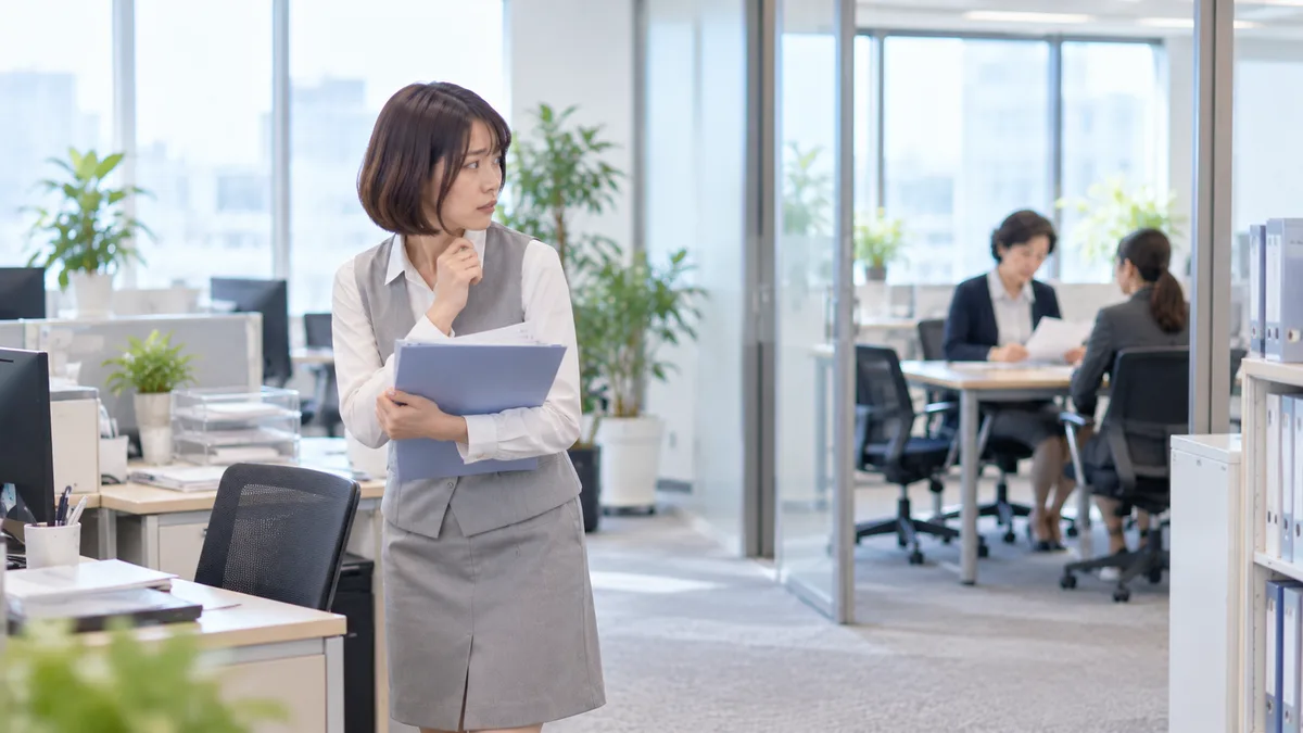 職場で高圧的な女性の様子を気にしながら、質問するための資料を持ったまま話しかけることをためらう女性。