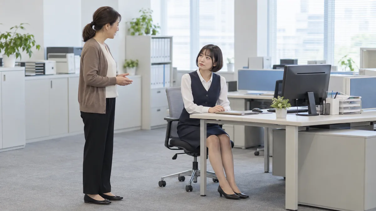 机に座り、立っている同僚へ顔を向ける女性