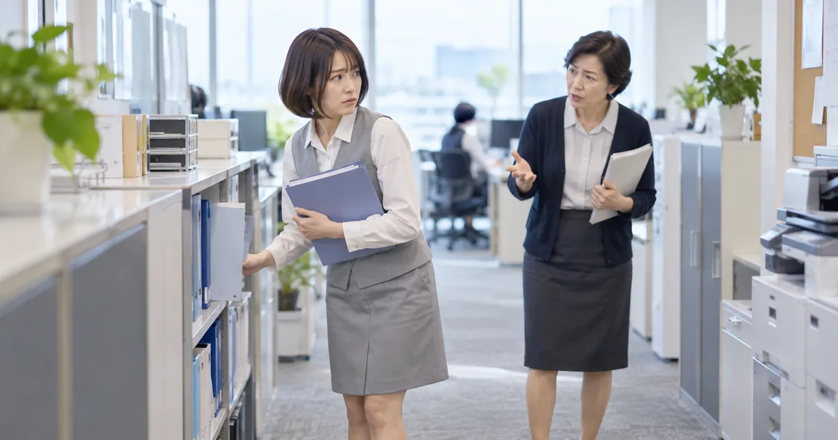 職場の書類棚の近くで50代女性から強い態度で仕事を指示され、戸惑いながら話を聞いている30代女性。