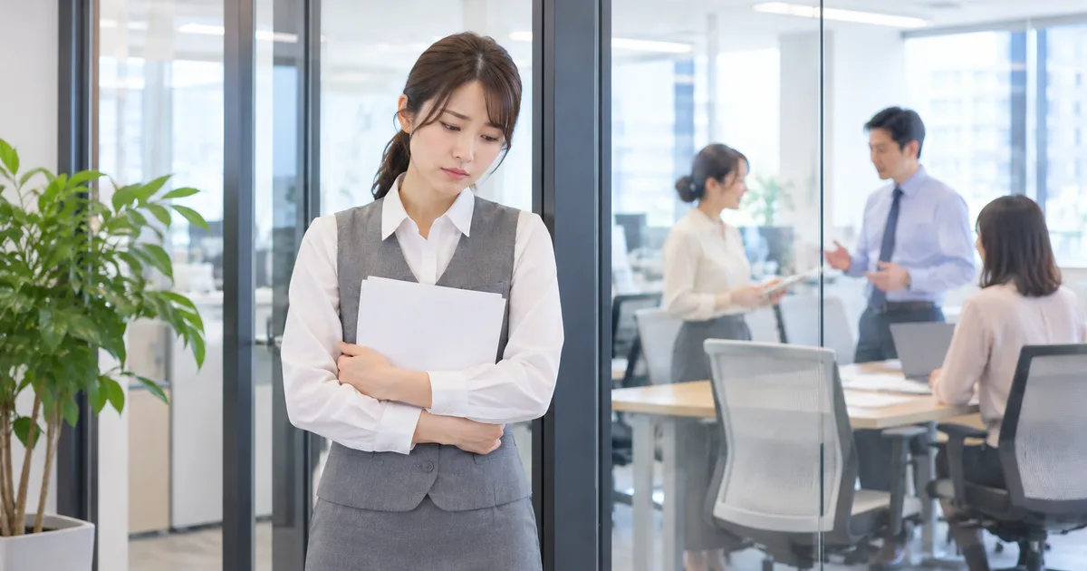 グレーの制服を着た女性が書類を抱え、ガラス仕切りの向こうで話す同僚たちから離れて立っている